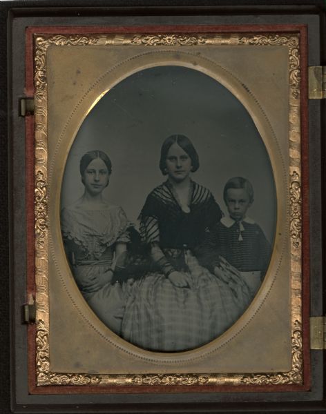 Sixth plate ambrotype of three-quarter length group portrait of two young women, sitting, and a young boy, standing, all facing forward. The woman on the left is wearing a striped fan-front dress with ruffles. The woman in the center is wearing a striped dress and black netting shawl pinned with a brooch. The boy is wearing a striped jacket with white collar and tasseled tie, and has his right hand on the lap of the center woman. Hand-coloring on cheeks. This image is on the left side of the case. 