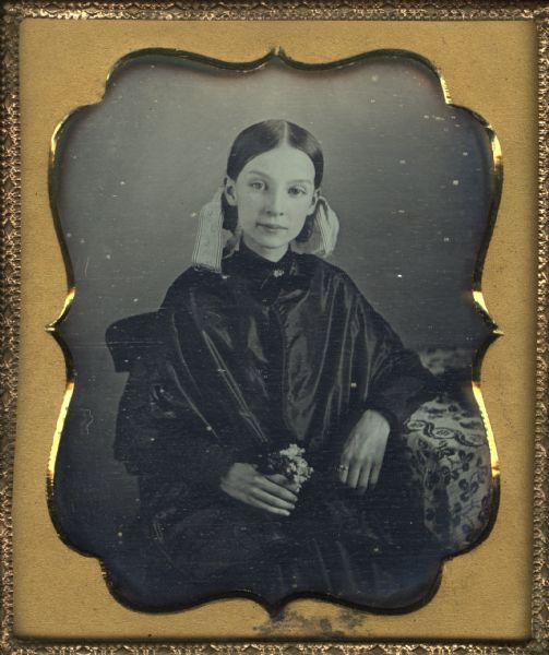 Sixth plate daguerreotype of Julia Smith. She is sitting with one arm resting on a cloth covered table and the other hand in her lap holding a small bouquet of flowers. Her hair is smoothed back behind her ears and tied up with wide ribbons, and she wears a ring on her left index finger. Julia Smith is the sister of Rose M. Bowman.