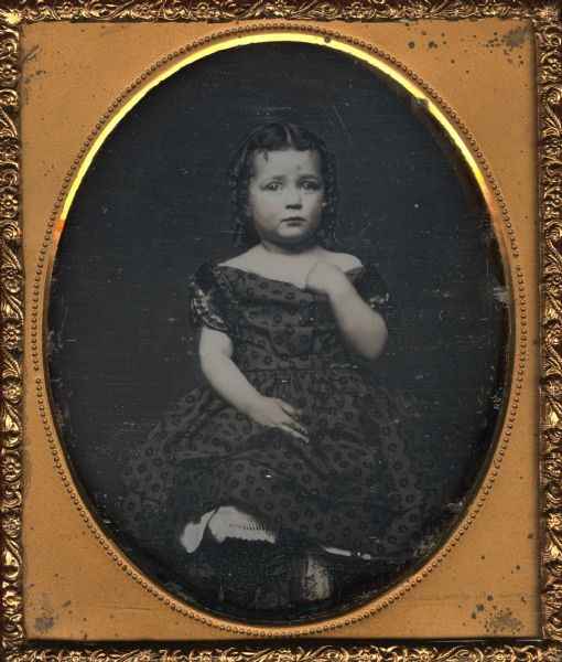 Sixth plate daguerreotype of Minnie Claire Donnel. The portrait is a full figure sitting, turned slightly to the left and looking front, left arm bent and left hand holding neckline of dress. Her hair is done in ringlets at the side. Hand-coloring on the cheeks.