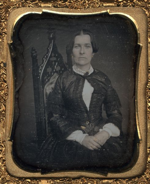 Sixth plate daguerreotype of Elizabeth Waddy Noyes, wife of Charles Cowper Noyes and mother of William Brayton Noyes. Waist-up portrait. She is sitting sideways in a Gothic-style chair with upholstered back, and has her hands in her lap. Hand-coloring on cheeks, gold details on jewelry including earrings, a collar pin, rings on her fingers and on the belt at her waist.