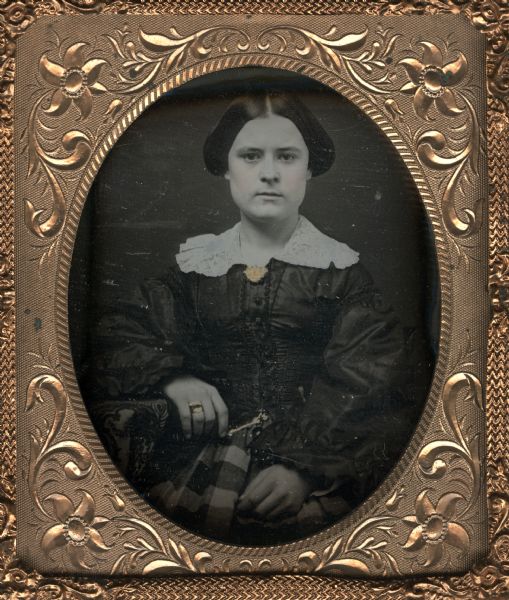 Sixth plate daguerreotype of Louise Noyes, sister of William B. Noyes. The portrait is a half figure, seated subject looking directly front, right elbow resting on cloth covered surface, wearing a lace collar and striped skirt. Hand-coloring on cheeks, and gold details on her jewelry including collar pin, ring on her finger and on the belt at her waist.