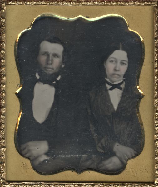Sixth plate daguerreotype of William and Emma Godfrey. The couple is shown waist-up seated next to each other with their hands crossed on their laps. There is hand-coloring on the cheeks.