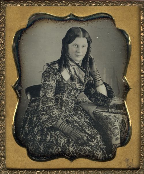 Sixth plate daguerreotype of Rosellah Smith (Donnel) Bowman. Mrs, Bowman was married to Samuel Hunter Donnel, architect, from 1854 to 1860. Samuel and Rosellah had one daughter Minnie or Mina. She married John Bowman 3 years after Donnel's death. This portrait was taken either before or at the time of her marriage to Samuel. The portrait is a half figure wearing floral print dress, shown facing front, looking off-center to the right. Her bent left elbow is resting on a cloth covered table with books, with her left hand resting on her left ear. She is wearing fingerless gloves. Hand-coloring on cheeks.