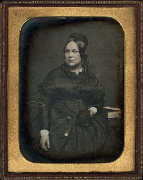 Quarter plate daguerreotype of Elizabeth S. Jaques Upham (1815-1883), wife of Don A.J. Upham, Milwaukee Lawyer. The portrait is a three-fourths figure, head front and turned slightly to the left. She is sitting near a table holding a bible. She is wearing a dark dress with a collar pin, and has a watch attached to a cord hanging from her waist.