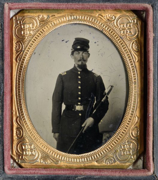 Sixth plate ferrotype/tintype of a Civil War Soldier. Three-quarter length portrait, perhaps Captain Edward Devlin, of Mineral Point, holding a sword. He served in Company E, of the 30th Wisconsin Volunteer Infantry. Hand-coloring on cheeks, and gold details on buttons and epaulets of military uniform.
