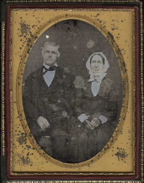 Quarter plate daguerreotype of Donnel and wife. Full figures, seated. Mr. Donnel wears a coat, vest, and tie, and has his left arm behind his wife. Mrs. Donnel has her hands in her lap and is wearing fingerless gloves. She is wearing a white bonnet, a white bow at her neck, and white cuffs. Hand-coloring on cheeks and lips. They were the parents of Samuel Hunter Donnel of Sandusky, Ohio, and the in-laws of Mrs. Rose M. (Donnel) Bowman. 