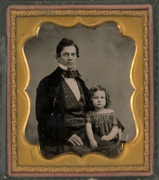 Sixth plate ambrotype of Edward P. Bridgman and his daughter in Madison, WI. Seated half figure of man facing front, torso turned to right, wearing coat and printed vest, standing collar and bow tie, with arm around small girl. Girl standing on man's left with hands on his left leg, in printed dress, with curled hair. Hand-coloring on cheeks and lips of both figures. 