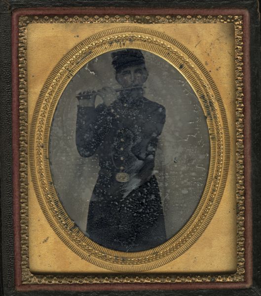Sixth plate ferrotype/tintype of a Civil War soldier standing while playing a fife. He is facing front, dressed in his uniform long coat, buttoned up, belt buckled, and with cap on. Hand-coloring on cheeks, and on gold details on buttons and belt buckle.