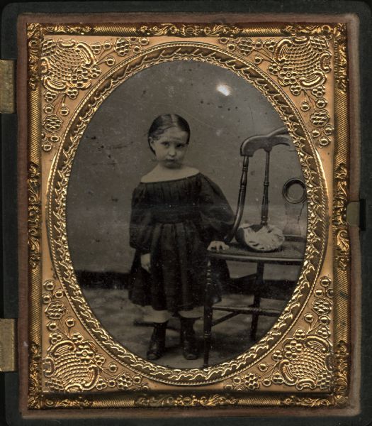 Sixth plate ferrotype/tintype of female child. Full figure facing front, standing with hand on chair, wearing belted, pleated shortwaisted dress off both shoulders, with long puffed sleeves, over tubular pantalettes (or "trowsers") and boots. Hair is pulled back and holds ornament on temple. Hand-coloring on cheeks. Object on chair is possibly a bonnet. 