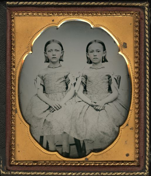 Sixth plate ambrotype of Tillie (Feb. 22, 1849-1864) and Anna Matilda Baker, possibly twins. Seated full figures with matching off-the-shoulder dresses, necklaces, and earrings, with hands in laps. Hand-coloring on cheeks and gold details on earrings and necklaces. Girl on left has light eyes, girl on right has dark eyes. 