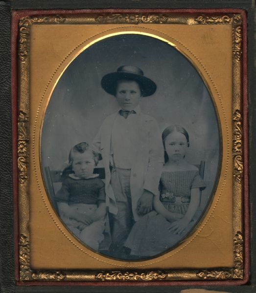 Sixth plate ferrotype/tintype of Bob La Follette with his brother William and sister Josephine. Bob and Josephine are seated; William is standing between them, all facing front. Bob wears pants, and a short sleeve shirt with white collar. William wears a white long coat, necktie, suspenders, and dark brimmed hat. Josephine wears a short sleeved dress, off-the-shoulder with quilted bodice, and a belt into which three flowers are tucked. Hand-coloring on cheeks. 