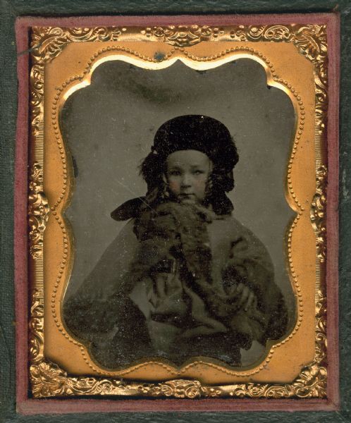 Ninth plate ambrotype of Horace A.J. Upham (1853-1919) as a child. Seated half figure facing front, wearing fur-trimmed overcoat and hat. Side curls appearing underneath hat. Hand-coloring on cheeks.
