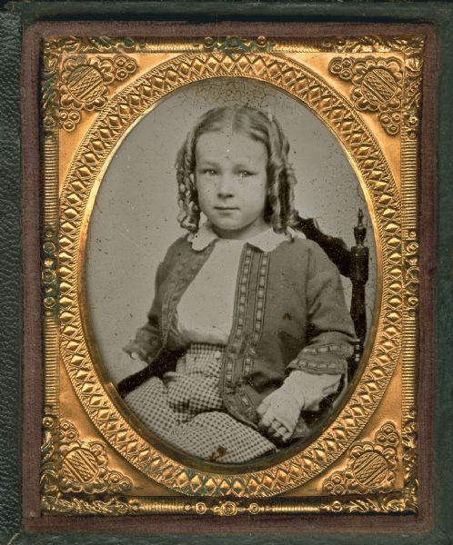 Ninth plate ambrotype of Horace A.J. Upham (1853-1919) as a child. Seated half figure facing front, torso turned to left, wearing jacket, shirt, checked pants, and sausage curls. Hand-coloring on cheeks. 