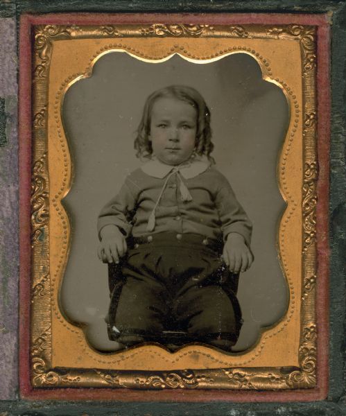 Ninth plate ambrotype of Horace Upham (1853-1919) as a child. Seated three-quarter length figure facing front, wearing sausage curls, knickers with brass buttons at waist, and jacket with white collar, tasseled tie, and brass buttons. Hands rest on chair. Hand-coloring on cheeks. 