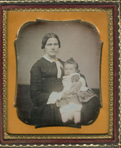 Sixth plate daguerreotype of Jane Sweeney Oakley with infant George Walter Oakley. Seated half figure facing front, torso to right, wearing dress with white collar and lace-edged scarf tied with brooch, drop earrings, and holding infant with left arm. Hand-coloring on cheeks and gold details on brooch. 