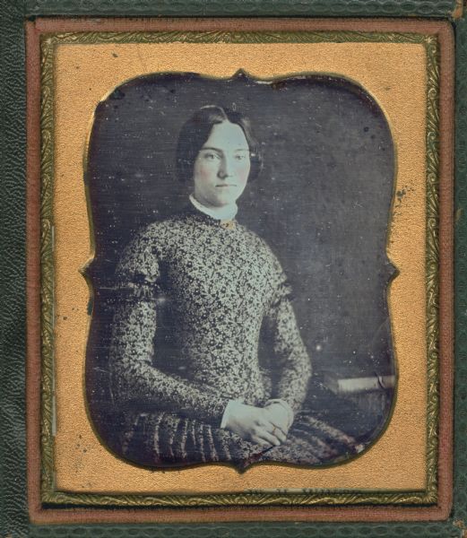 Sixth plate daguerreotype of Harriet Sweeney Hough. Seated half figure facing front and to the right, wearing long-sleeved printed dress with small white lace collar and brooch at throat, with hands folded in lap. A closed book lies on a table on the right. Hand-coloring on cheeks and gold details on brooch.