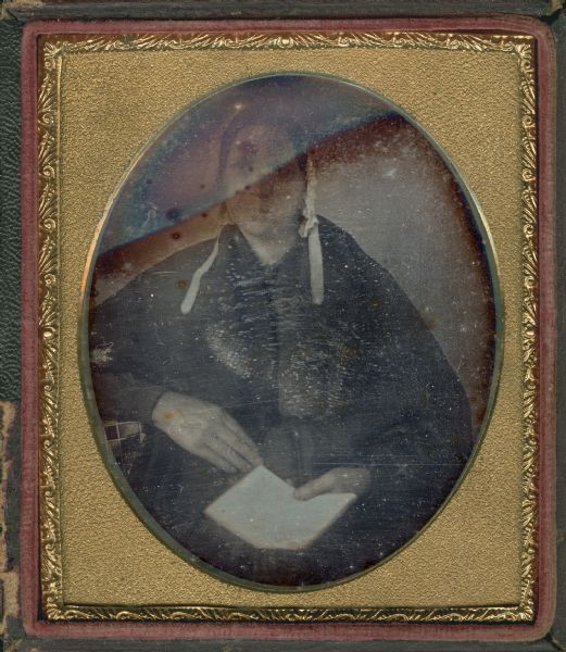 Sixth plate daguerreotype of unidentified woman, probably Mary Oakley Hough. Seated half figure facing front and gazing left, wearing untied bonnet, with right elbow resting on a what may be a cloth-covered table. In her left hand she is holding an open book in her lap. 