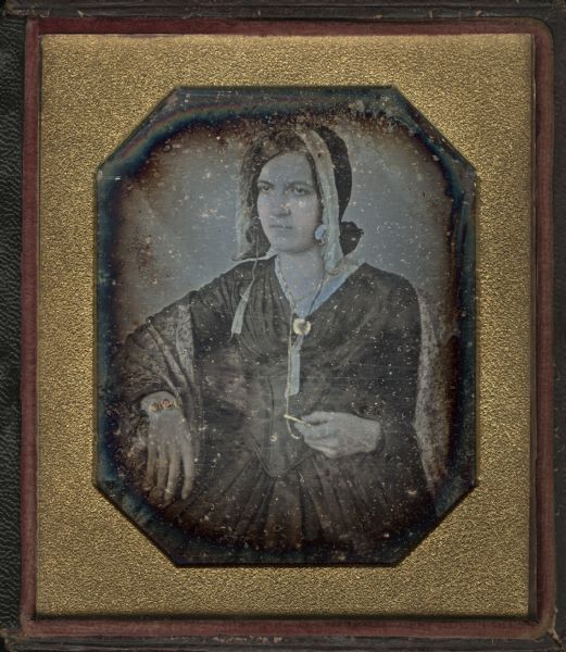 Sixth plate daguerreotype of an unidentified woman, probably Mary Oakley Hough. Half figure facing front and slightly left, with right elbow resting on a surface, and left hand holding one end of watch chain. She is a wearing a bonnet, shawl, and long-sleeved dress. Hand-coloring on cheeks and gold details on brooch, watch chain, and bracelet.