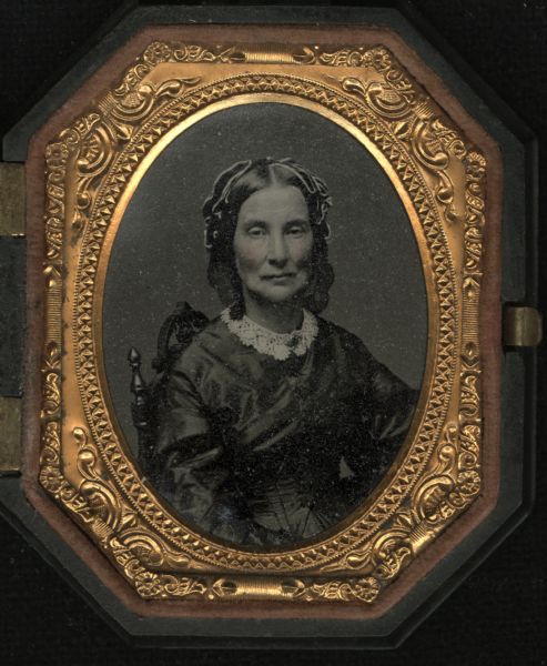 Ninth plate ferrotype/tintype of unidentified woman. Waist-up seated portrait, facing front. She is wearing a lace collar with brooch, a long-sleeved dress that appears to be silk or satin, and silk or satin hair ribbons. Hand-coloring on cheeks. 