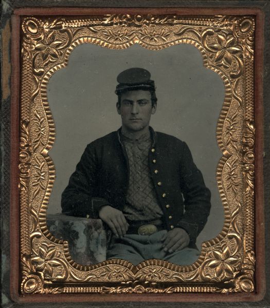 Sixth plate ferrotype/tintype of James Hurd. Seated half figure with arm resting on cloth-covered table, wearing Union Army coat and cap, check patterned shirt, blue tinted pants, and US belt buckle. Hand-coloring on cheeks and pants, and gold details on buckle and coat buttons. 