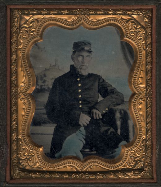 Sixth plate ferrotype/tintype of James W. Gorum in front of a painted backdrop. Seated three-quarter figure with beard and mustache, facing front with elbow on cloth-covered table, wearing Civil War Union coat, cap, and blue-tinted pants. Hand-coloring on backdrop, cheeks, trousers, cap, and gold buttons. Gorum served in Company G, 1st US Sharpshooters (Berdan's Sharpshooters). 