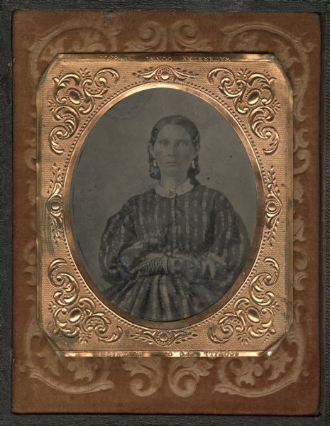 Three-quarter length ninth plate ferrotype/tintype of Mrs. Larsen, wife of Peter Larsen. She is wearing a striped, long-sleeved dress with white collar, lace gloves, a brooch at her neck, and drop earrings. Her arms are folded. Hand-coloring on cheeks, and gold details on jewelry. Mounted on the inside cover of the case, with Peter Larsen on the right side. 