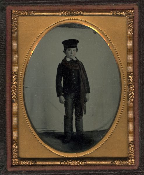 Ninth plate ambrotype. Full-length portrait of Henry A. Cooper, as a young boy, standing in front of a backdrop. He is wearing a coat, scarf, hat, and what may be knee-length boots. Hand-coloring on cheeks.