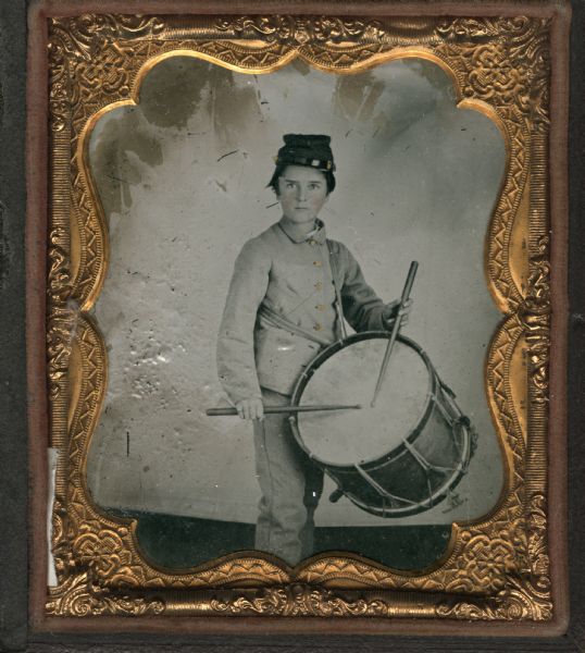 Ninth plate ferrotype/tintype.  Three-quarter length portrait of Henry A. Cooper, as a young boy, standing in front of a backdrop. He is wearing a military uniform, and is holding drumsticks over a drum slung over his left shoulder. Hand-coloring on cheeks and gold details on buttons.