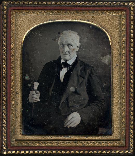 Sixth-plate daguerreotype. Waist-up portrait of an unknown man sitting in a chair. He is holding a cane in one hand, and is wearing a suit coat, vest, and necktie. Hand-coloring on cheeks.