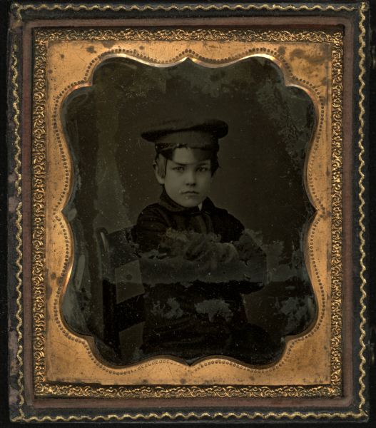 Sixth-plate ambrotype. Waist-up portrait of James D. Butler, Jr. sitting in a chair. He has his arms crossed over his chest, and is wearing a jacket and a wheel hat.