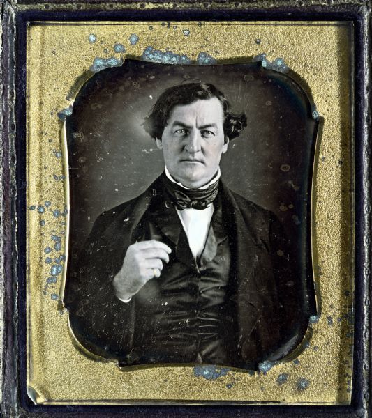 Sixth plate daguerreotype of unidentified man. Bust portrait facing forward, wearing suit, cravat, and stand collar, with right hand holding clothing near lapel. A gold ring is on his little finger. Hand-coloring on cheeks and ring. 