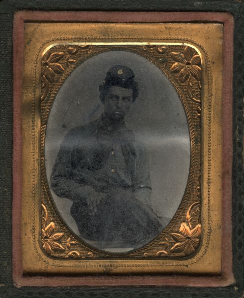 Ninth plate ferrotype/tintype of Marcus S. Pratt. Served in Company G. 15th Regiment, Wisconsin. He is sitting in a chair with his knee propped up, with one hand on his shin and the other on his thigh. He wears a large, floppy tie. Gold details on cap and buttons.
