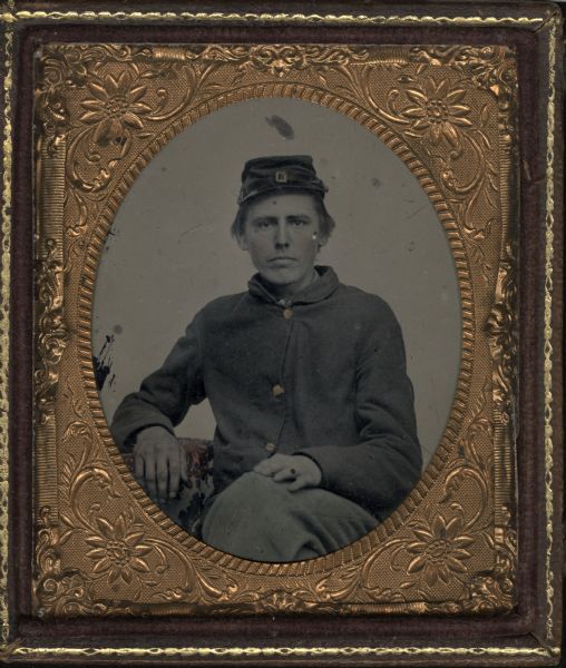 Sixth plate ferrotype/tintype. Three-quarter length portrait of James K. Newton sitting with one leg crossed over the other, one hand resting on his thigh, and the other arm resting on a cloth-covered table. Hand-coloring on cheeks and gold details on cap and buttons.