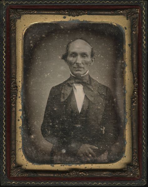 Quarter plate daguerreotype waist-up portrait of unidentified middle-aged man with receding hairline with his hands folded toghether at his waist. He is wearing a coat with wide lapels, vest, white shirt with stand collar, and dark cravat with long tails.