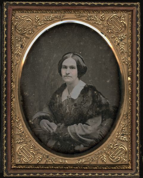 Quarter plate daguerreotype of unidentified woman. Half figure facing front and slightly left. She is wearing a white collar, bell sleeves, and fur cape and cuffs, and a collar pin. She is resting one arm on a table, clasping her hands together. Hand-coloring on cheeks. 