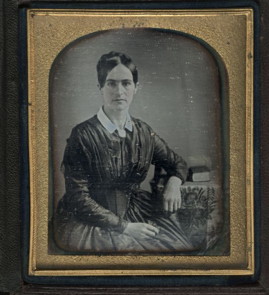 Sixth-plate daguerreotype. Three-quarter length portrait of an unidentified woman, facing front. She is sitting in a chair, with one hand in her lap, and the other arm resting on a cloth-covered table, on which a book is resting. Hand-coloring on cheeks.