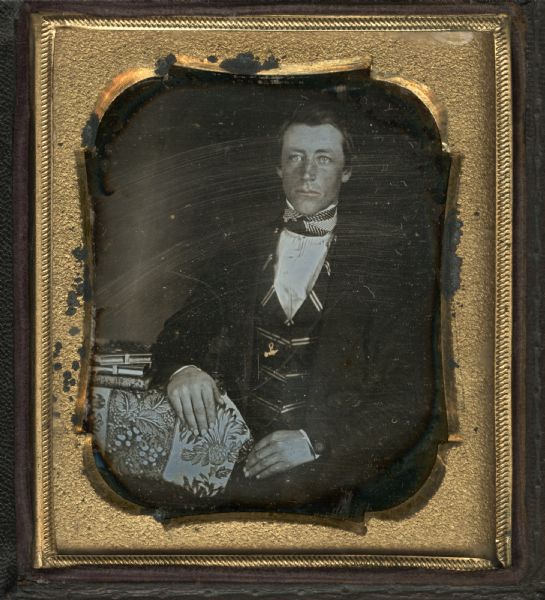 Sixth-plate daguerreotype. Waist-up portrait of unidentified man who is wearing a suit coat, striped vest, and a cravat. He is sitting in a chair with one hand in his lap and the other arm resting on a cloth-covered table on which is sitting a stack of books. Hand-coloring on lips and gold detail on watch chain.