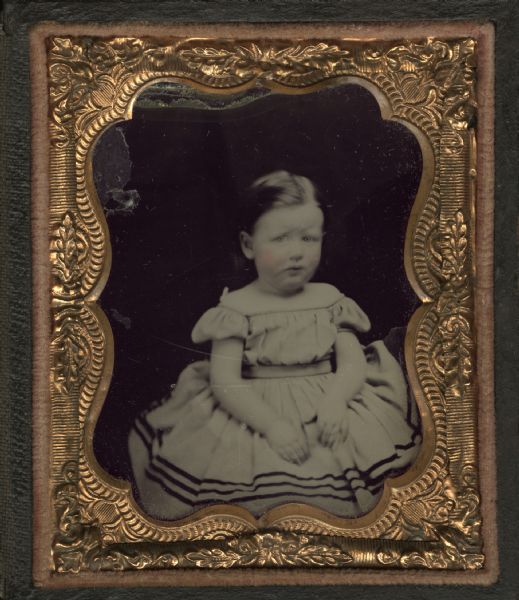 Ninth plate plate ambrotype. The child is sitting, wearing an off-the-shoulder dress belted at the waist. Dark trim along hem. Hand-coloring on cheeks.