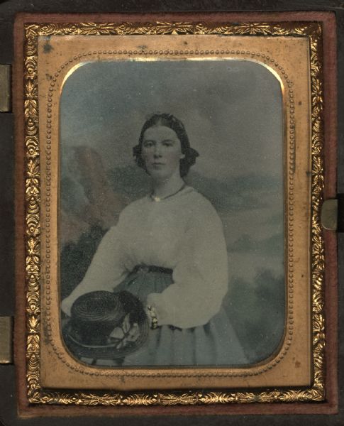 Ninth plate ferrotype/tintype. Three-quarter length portrait of a woman sitting in front of a painted backdrop. She is holding a straw hat in her lap. Hand-coloring on cheeks and lips, and gold details on necklace and bracelet.