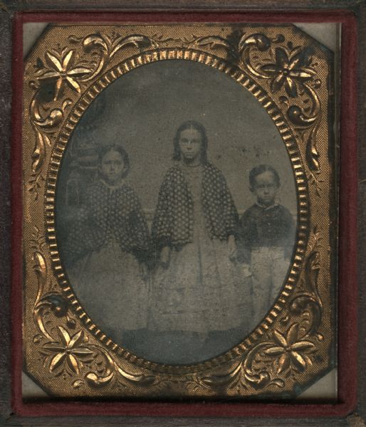 Sixth-plate ferrotype/tintype. Group portrait of three children standing in front of a painted backdrop. The two young girls are wearing matching jackets. Allegedly the nieces and nephew of Franklin B. Howard.