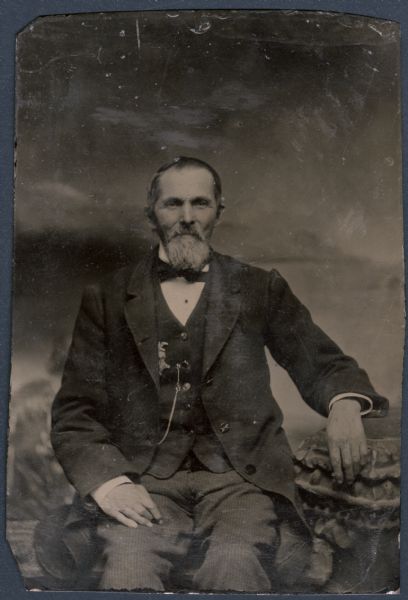 Sixth plate ferrotype/tintype. Three-quarter length portrait, facing forward, of Truman Bartlett, from Stevens Point, Wisconsin, sitting on a bench in front of a painted backdrop. He has one forearm resting on a stone column, and the other hand resting on his thigh. He is wearing a suit coat, vest, necktie, and watch chain. A badge on his vest is slightly covered over with his suit coat. He has a moustache and a beard. Hand-coloring on cheeks.