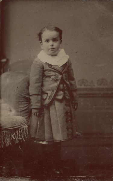 Ferrotype/tintype of John C. Hawley, age 6, Evergreens, Madison, Wisconsin. He is wearing a white lace collar, embroidered jacket, vest, and skirt, and is stands against an upholstered settee, facing forward.