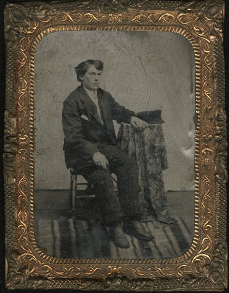 Quarter plate ferrotype/tintype of Ceylon Childs Lincoln. Full-length seated pose facing 45 degrees to the right, with left arm on cloth covered table and right hand on knee. His cap sits on the table, and under his feet is a striped rug. 
Born April 18, 1850 in Naperville, IL. In 1856 he moved to Wautoma, WI and in 1863, at the age of 14, enlisted in the Union Army (35th Wisconsin Volunteer Infantry--Company D) until Dec. 1, 1865. He was later employed as a printer and wrote "Personal Experiences of a Wisconsin  Raftsman" (see Wisconsin Historical Society Proceedings, 1910, p. 180).  He married Melinda J. Duncan of Richford in 1870; became an early settler of Tomahawk in 1887; and in 1895 was appointed a janitorship in the Museum of the State Historical Society and served until 1909.