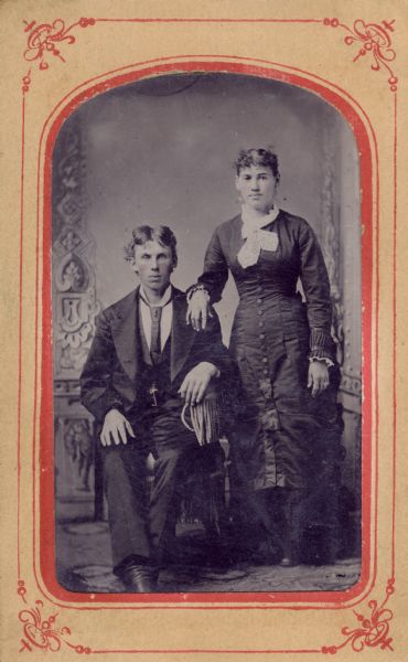 Full-length ferrotype/tintype of an unknown man, and Esther Lomas from Lancaster, Wisconsin, in front of a painted backdrop. The man is seated, and the woman is standing at his left with her right hand on his shoulder. Hand-coloring on cheeks, and gold details on man's tie pin, woman's earrings, rings, and collar pin.