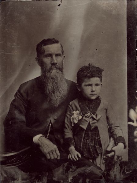 Three-quarter length ferrotype/tintype of a seated man, thought to be Louis Luce, and standing next to him, his daughter Louise, from Stevens Point, WI. His arm is around the girl standing at his side, and his elbow is resting on a table. He has a chest-length beard and is wearing a coat with the top buttons closed, and a watch chain hanging beneath. The girl is wearing an overcoat with satin bow at the collar and a bow pinned on the left, and her right hand is resting on the man's thigh. 