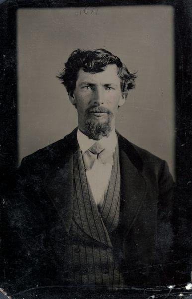 Three-quarter length ferrotype/tintype of Cyrus Merryfield from Stevens Point, Wisconsin. He faces front and is wearing a three-piece suit with striped vest and tie, and has a mustache and goatee. Hand-coloring on the cheeks. 