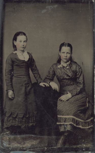 Full-length ferrotype/tintype of Nellie Orms and Hattie Beers, Sunday school pupils of Lillian Luce Holt. The girl on the left is standing with her hand on a covered table, and is wearing a solid color dress, white collar and locket. The girl on the right is seated with her forearm resting on the same table, and is wearing a printed dress and necktie. Cheeks are hand-colored.  