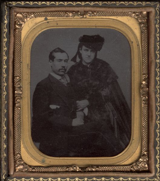 Sixth plate ferrotype/tintype of John Purcell and his wife. He is sitting facing halfway to the right, with his hands folded in his lap with his leg crossed. She is standing, facing front and leaning into him with her hand on his arm. She is wearing a fur cape, cuffs, and a hat. He is wearing a suit and has a moustache. 