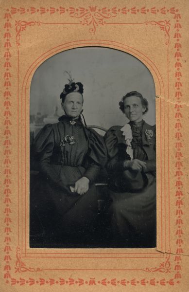 Ferrotype/tintype of Mary E. Stewart and Jennie E. Woodworth at Brenham(?), Texas, 1889. Miss Stewart was a Milwaukee educator and journalist. Both are seated, facing front, with hands folded on laps. The left figure is wearing a hat tied under her chin, a collar pin, and a rose pinned to the front of her dress. The right figure is wearing gloves, a white lace ruffle on the left side of her jacket, a collar pin, a rose pinned to her lapel, and a hat in her lap.