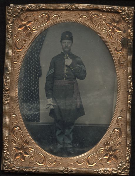 Quarter plate ferrotype/tintype of unidentified Civil War Union officer. Full-length portrait of man standing facing front, with his hand inside his uniform coat, and the other hand at his side holding his gloves. He us wearing a cap, red sash, blue trousers, and rank chevrons on his sleeves. Cap insignia, cheeks, gold buttons, sash, chevrons, and trousers are hand-colored. Draperies are part of the backdrop. 